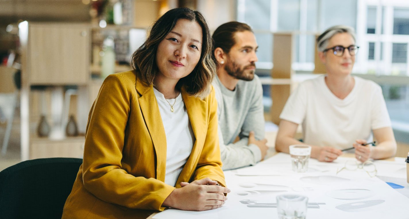 A smiling Asian woman wearing a yellow blazer sits beside a couple of her professional colleagues working together on a project