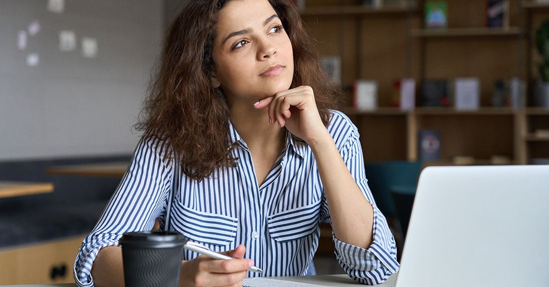 A young professional woman wearing a striped button down shirt holds a pen at her desk in front of a laptop, staring off into the distance, thinking.