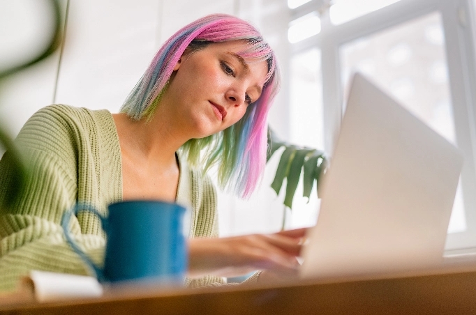 A young woman with pink hair works on a laptop
