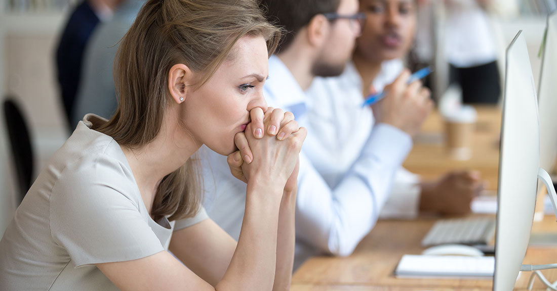 A woman sits at a table in an office meeting in front of a computer leaning her chin on her folded hands looking concerned