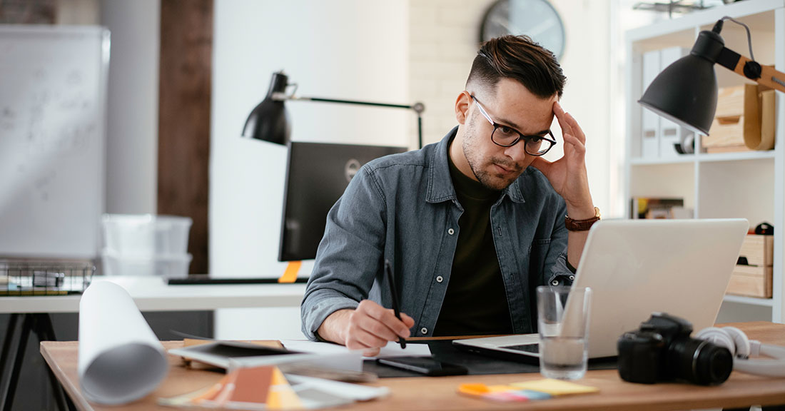 Man wearing glasses sitting at desk in an office setting looking concerned while using a laptop.