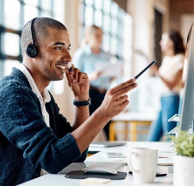 A smiling man working in customer support points at his computer screen with a pen.