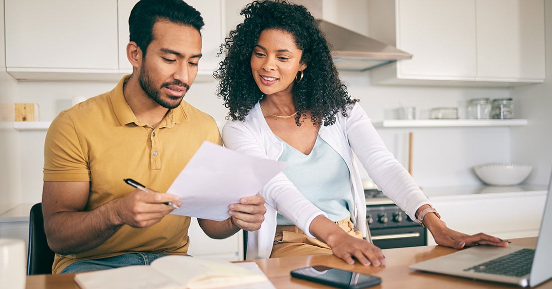 A man and woman sit at a kitchen counter reviewing student loan documents and researching SAVE Plan updates online.