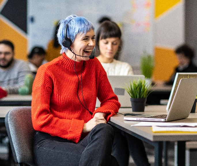 Woman with blue hair and red sweater laughing in front of a computer and wearing a headset