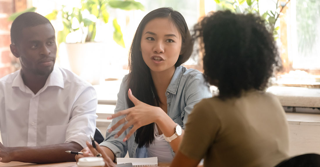 Three financial aid professionals in a college office discuss cohort default rates at a table while reviewing documents over coffee. 