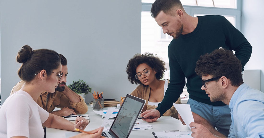 A diverse group of serious-looking young professionals meet around a table in a modern office setting.